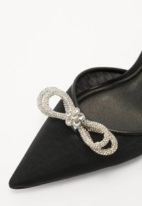 Chaussure noire à bout pointu en tissu texturé, ornée d'un nœud en strass scintillant et d'un accent floral en argent.