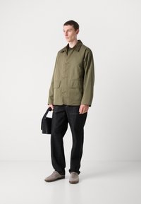 Olive green, lightweight jacket with a collared neck, front button closure, and two large patch pockets paired with dark denim trousers and gray sneakers.