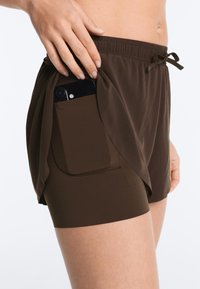 Hand placing smartphone into side pocket of brown athletic shorts with inner compression layer and outer loosened layer.