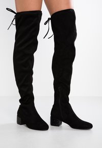 Botas negras de ante sobre la rodilla con punta afilada, tacón cuadrado con detalle de purpurina y cierres ajustables con cordones en la parte superior.