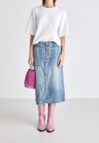 Jupe en denim avec un devant à boutons et deux poches, portée avec un haut blanc à manches courtes côtelé et des bottes roses jusqu'au genou, tenant un sac à franges rose.