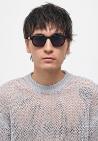 Pyöreät mustat aurinkolasit paksulla kehykellä. Mallilla on päällään vaaleanharmaa, löyhästi kudottu neulepaita, jossa on ribattua kaula-aukkoa ja huomattavaa tekstuuria.