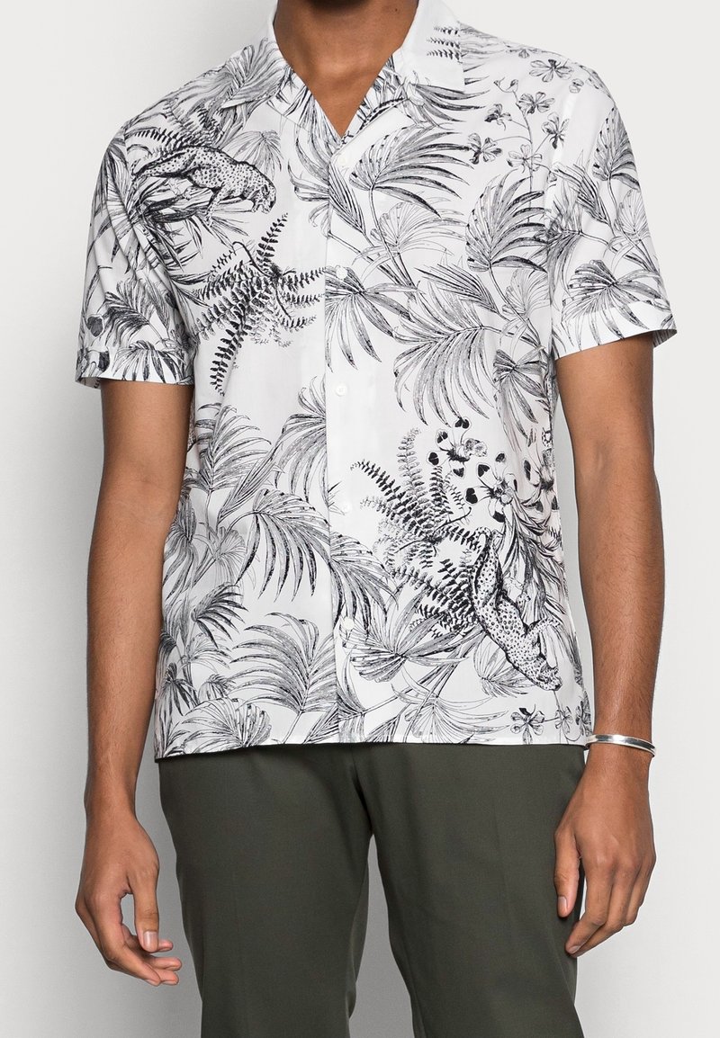 Chemise blanche à manches courtes boutonnée avec imprimé botanique noir et léopard portée par une personne portant un pantalon vert olive.
