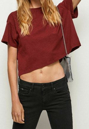 Vrouw met een cropped bordeauxrode T-shirt en zwarte jeans, met een zilveren kettingtas die aan haar rechterzijde hangt.