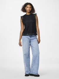 Tânără cu păr creț purtând un top negru fără mâneci din dantelă, blugi largi de culoare albastru deschis și pantofi negri, stând în picioare pe un fundal alb.