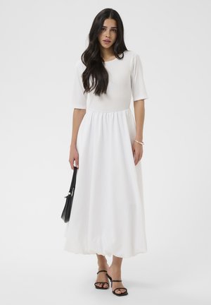 Frau mit langen dunklen Haaren, die ein weißes Midikleid mit Ellenbogenärmeln trägt, hält eine schwarze Handtasche und trägt schwarze Riemchenabsätze.