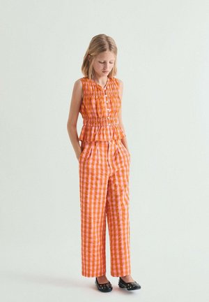 Niña joven vistiendo un top sin mangas de cuadros naranja y rosa junto con pantalones a juego, de pie con las manos en los bolsillos y mirando hacia abajo.