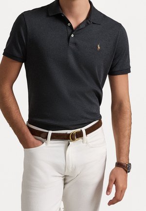 Homme portant un polo gris foncé avec un petit logo brodé, un pantalon blanc, une ceinture marron avec une boucle dorée et une montre-bracelet avec un bracelet marron.