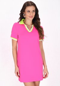 Neonpinkin mekko, jossa on V-pääntie, lyhyet hihat ja vastakkaiset limevihreät kaulus ja hihojen reunat. Sileä pinta, rento muotoilu.