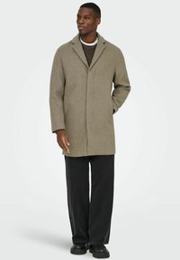 Olive roheline villane mantel, millel on sälguline krae, ühe nupuga disain ja küljetaskud, koos mustade laia jala pükstega ja mustade kingadega.