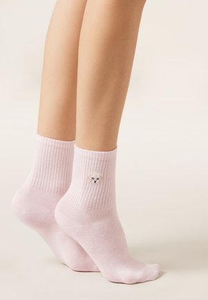 Calzini rosa chiaro alla caviglia con un piccolo viso di cane bianco ricamato sul lato esterno, indossati a piedi nudi su uno sfondo semplice.