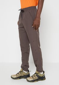 Ciemnobrązowe, lekkie spodnie z elastycznym pasem i sznurkiem do regulacji. Posiada boczne kieszenie i zwężane nogawki. Nosi je w połączeniu z beżowymi butami turystycznymi.