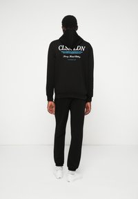 Czarna bluza z kapturem z nadrukowanym tekstem na plecach, w połączeniu z dopasowanymi czarnymi dresami i białymi sneakersami. Ubranie charakteryzuje się luźnym krojem.