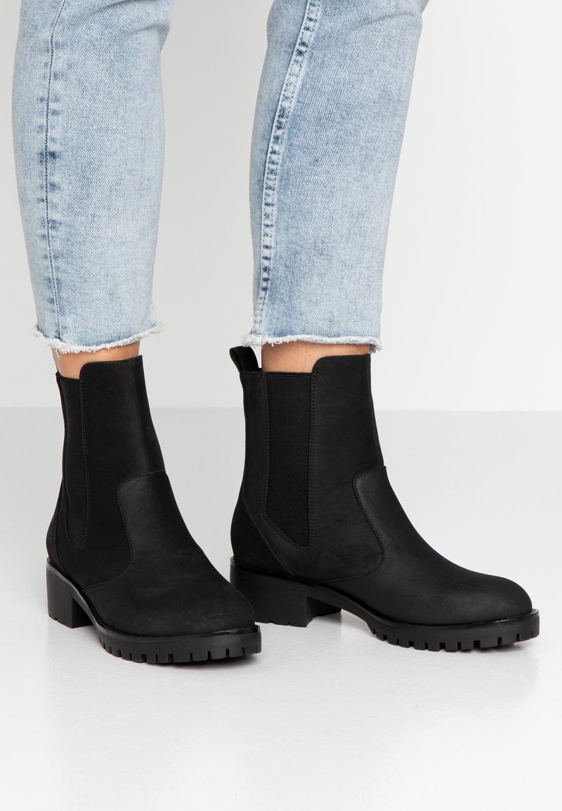 Personne portant un jean bleu clair et des bottines noires à talons épais et panneaux latéraux élastiques, debout sur un sol blanc.
