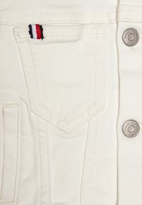 Bílá džínová látka s viditelným prošíváním, patková kapsa s červeným, bílým a černým pruhovaným akcentem a kovový knoflík s brandem "Tommy Hilfiger."