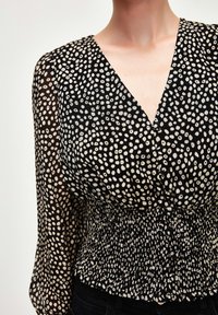 Blusa a fantasia bianco e nero con scollatura a V profonda, vita arricciata e maniche lunghe trasparenti; presenta forme tondeggianti e una texture di tessuto leggera.