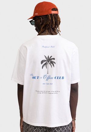 Mand iført hvid t-shirt med sort palme og blå tekst "The OUT of Office CLUB" og orange kasket, set bagfra mod en ensfarvet baggrund.