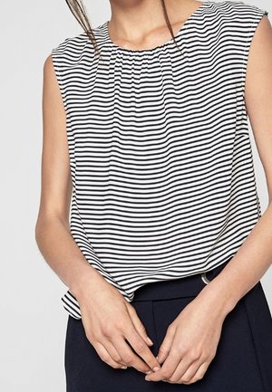 Frau trägt ein ärmelloses schwarz-weiß gestreiftes Top, das in dunkelblaue Hosen gesteckt ist, Hände locker an der Taille verschränkt.