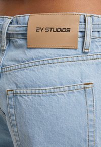 Světle modré džíny z denimového materiálu, zadní pohled ukazuje hnědou koženou nášivku s textem "2Y STUDIOS" nad pravou zadní kapsou.