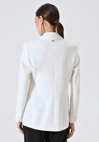 Witte op maat gemaakte blazer met lange mouwen en kraag. Beschikt over een achterste vent en een gouden logo accent bij de halslijn. Glad, gestructureerd stof.
