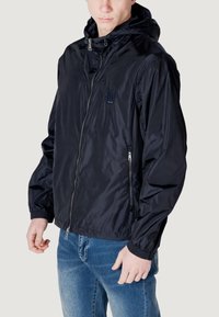 Giacca nera in nylon con zip e cappuccio, polsini elasticizzati e due tasche laterali. Presenta un piccolo logo sul petto e un design leggero.