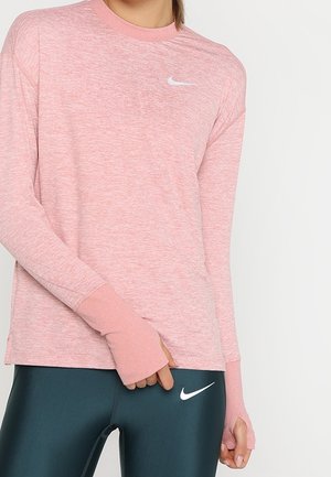 Kobieta ubrana w różową, długorękawową bluzkę z otworami na kciuki i ciemnozielone legginsy, obie części z białym logo Nike Swoosh.