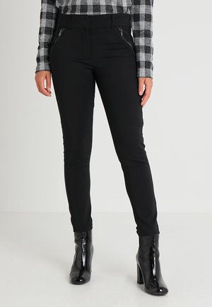 Frau trägt schwarze, eng anliegende Hose mit vorderen Reißverschlusstaschen, schwarze Stiefeletten aus Lackleder und ein grau-schwarz kariertes Langarmshirt.