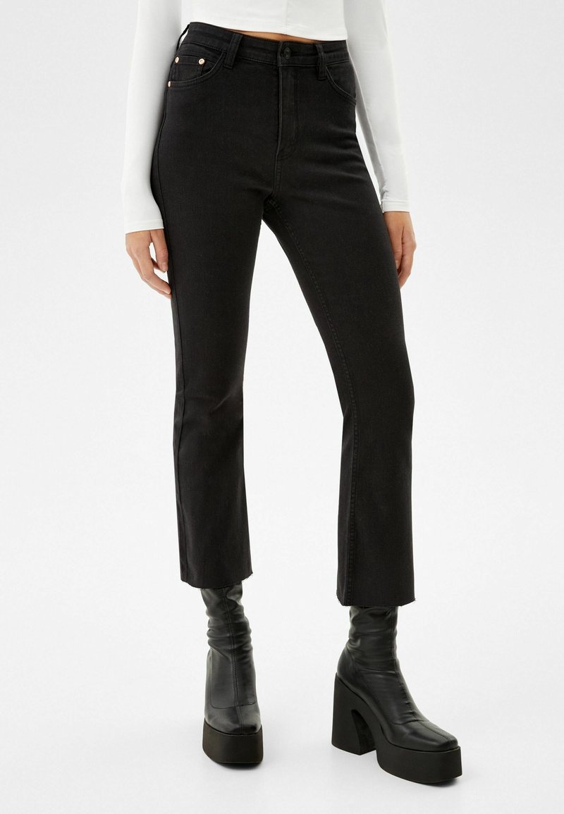 Personne portant un jean noir cropped taille haute, un top blanc à manches longues et des bottines noires à talons compensés, debout sur fond blanc.