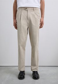 Man wearing beige pleated trousers, black loafers, and a white shirt standing on a gray floor against a plain light wall.