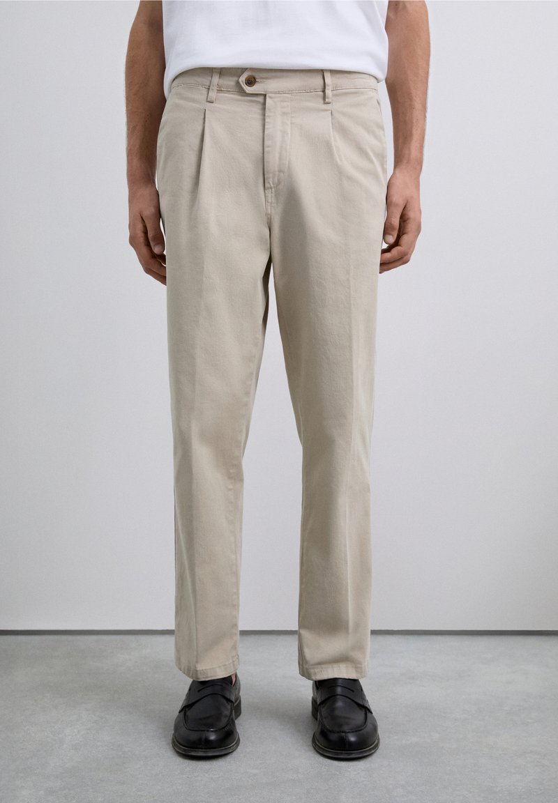 Man wearing beige pleated trousers, black loafers, and a white shirt standing on a gray floor against a plain light wall.
