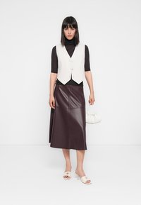 Chaleco blanco sobre un suéter de cuello alto negro, falda de cuero marrón, bolso blanco y sandalias de tacón blanco abiertas por delante. Texturas suaves, ajuste entallado.