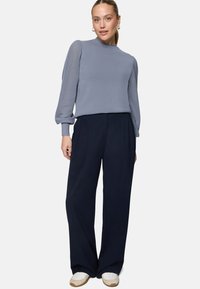 Femme portant un pull bleu clair avec des manches texturées, un pantalon large foncé, des baskets blanches, debout devant un fond uni.