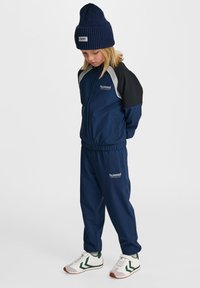 Marinblå kostym av slätt tyg med kontrasterande svarta och vita axelpaneler; matchad med en marinblå stickad mössa och vita sneakers med gröna detaljer.