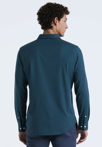 Teal langesleeve shirt met een button-down kraag, twee knopen bij de mouwen en een afgeronde zoom. Gemaakt van een gladde stof met een subtiele textuur.