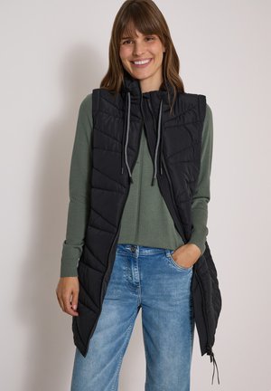 Femme souriante portant un gilet sans manches matelassé noir sur une chemise verte à manches longues et un jean bleu, debout avec une main dans la poche.