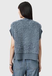 Gilet lavorato a maglia blu con texture fluffy sulla parte posteriore, maniche corte e orlo a costine. Indossato sopra jeans in denim a vita alta di colore blu chiaro.