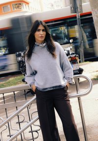 Pulover gri cu tiv dantelat, asortat cu pantaloni maro închis cu croială largă. Modelul stă lângă o balustradă metalică, cu elemente de fundal neclare.