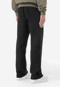 Pantalones de chándal negros con textura, presentan un corte holgado y una cintura elástica. Combinados con zapatillas blancas, mostrados desde atrás.