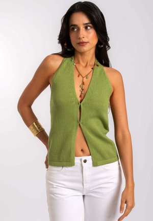 Top halter lavorato a maglia verde con scollatura a V profonda e apertura frontale, caratterizzato da una texture a coste. Abbinato a jeans bianchi e accessori dorati.