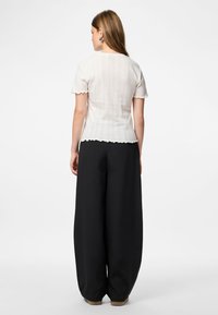 Ženska s ravnimi svetlo rjavimi lasmi nosi belo kratkoročno rebrasto majico in široke črne hlače, stoji ob običajni beli ozadju.