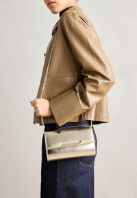 Metallic gold crossbody bag with a chain strap, smooth texture, rectangular shape, and a snap closure, worn against a tan leather jacket.