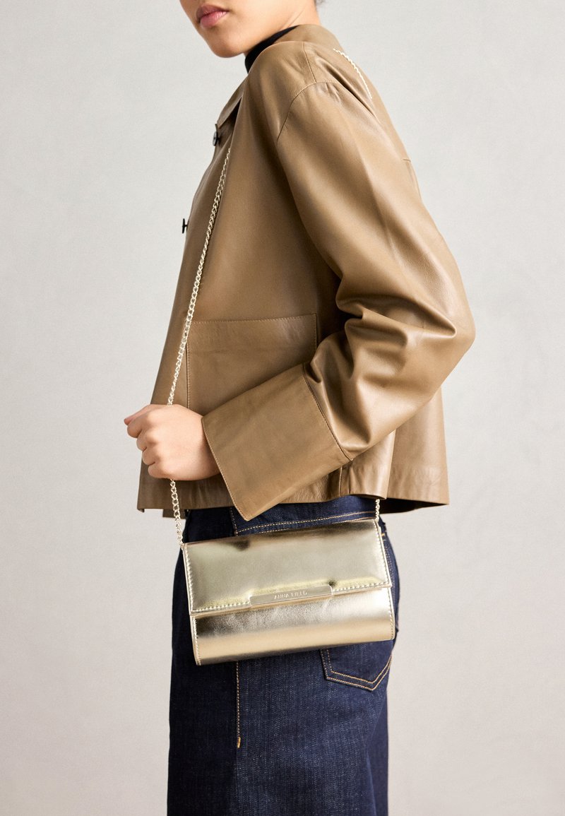 Metallic gold crossbody bag with a chain strap, smooth texture, rectangular shape, and a snap closure, worn against a tan leather jacket.