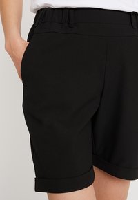 Persona che indossa pantaloni corti neri con orlo arrotolato e vita elastica, con la mano sinistra nella tasca e una maglietta bianca infilata visibile all'altezza della vita.