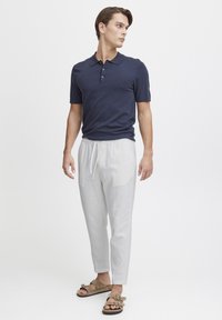 Marineblå kortermet polo, lysegrå tapered bukser med snøring, og brune sandaler med spenner, mot en enkel hvit bakgrunn.