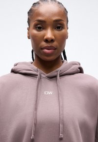 Εγγύς λήψη μιας γυναίκας με πλεκτά μαλλιά που φοράει μωβ φούτερ με κουκούλα και το λογότυπο "ICIW", κοιτάζοντας εμπρός με φόντο ένα απλό λευκό φόντο.