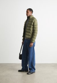 Olivengrøn pufferjakke med høj krave, parret med en mørkegrå sweater, bredbenede blå jeans og sorte sko; bærer en lille sort taske.
