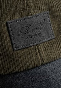 Olive green corduroy cap with a black felt patch featuring embossed text. Structured shape with contrasting textures and stitching details.