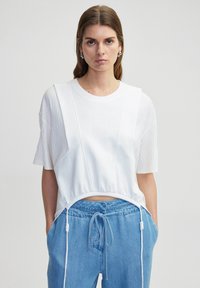 Bílý zkrácený tričko s texturovanými oversized rukávy a stahovacími okraji. V kombinaci s světle modrými kalhotami se stahovací šňůrkou. Hladká látka.
