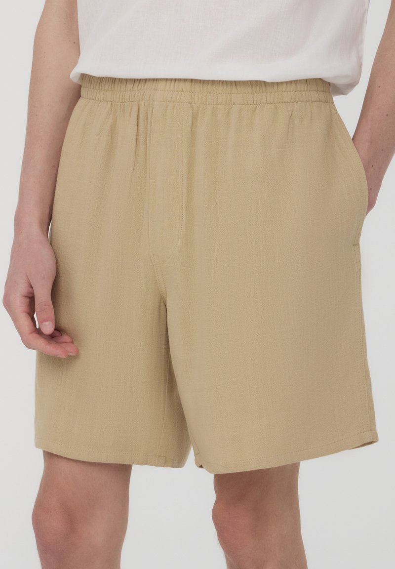 Pantaloni corti beige realizzati in un tessuto testurizzato, con vita elastica e tasche laterali, caratterizzati da un fit rilassato e design che arriva fino al ginocchio.