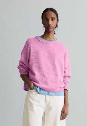 Frau trägt einen rosa strukturierten Pullover über einem blauen Hemd und cremefarbenen Hosen, steht mit einer Hand in der Tasche vor einem schlichten grauen Hintergrund.